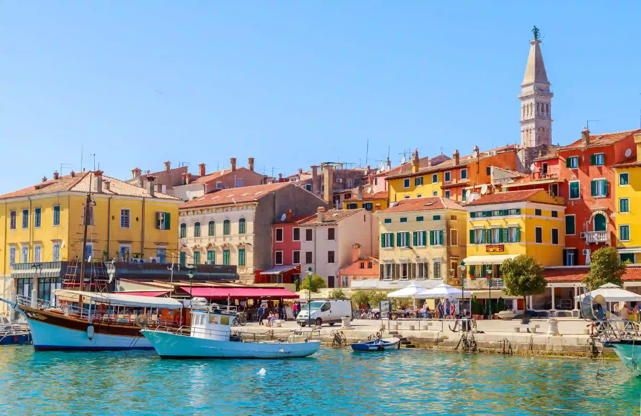 View of Rovinj from the water, showing boats docked at the harbour and colourful buildings. Slightly further back is a stone tower. The sky is blue and clear.