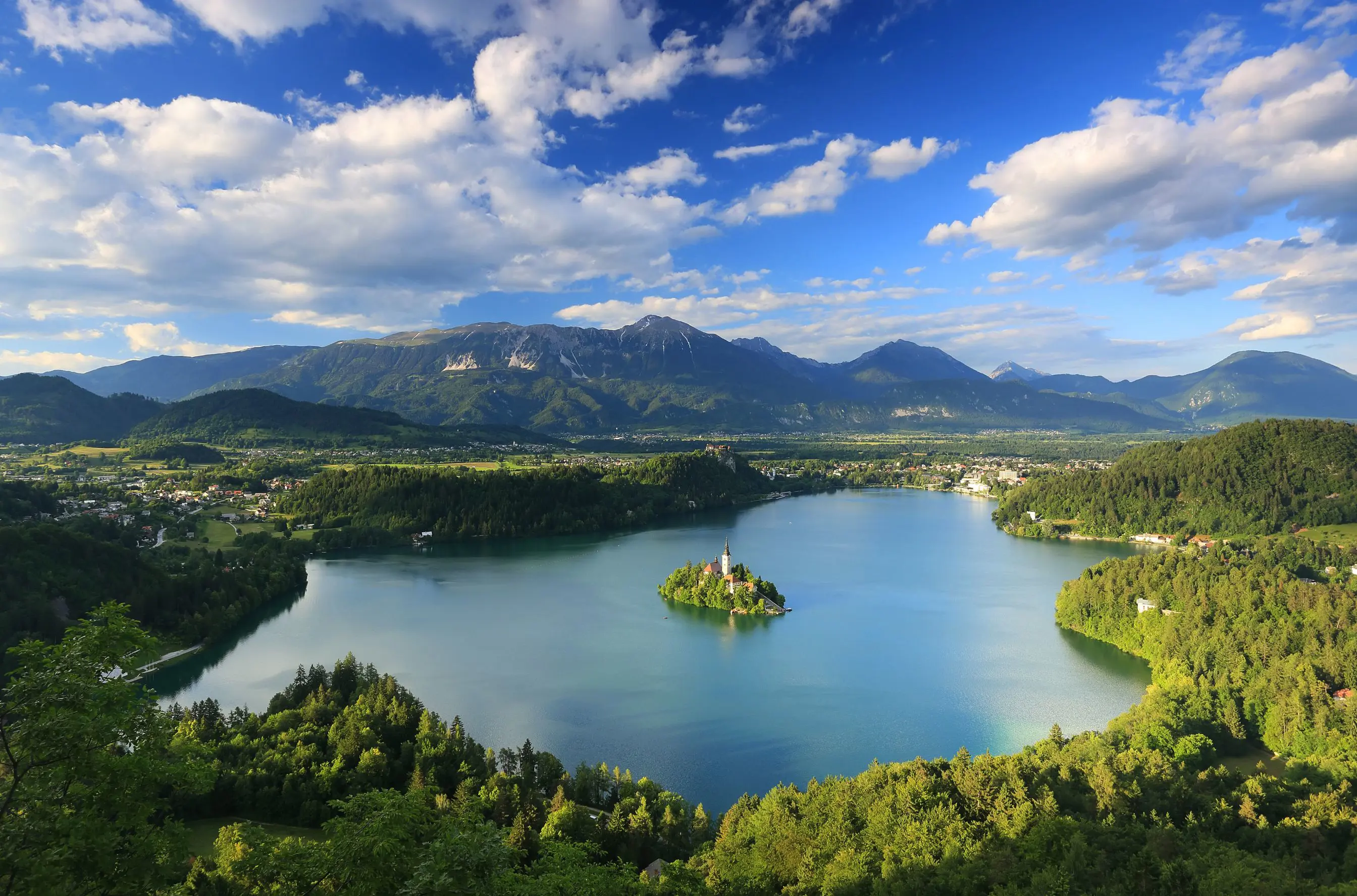  Wide angle shot of Lake Bled, showing its forested surrounding areas, and mountains in the distance. In the middle of the lake, there is a small piece of land with trees and a towered building.