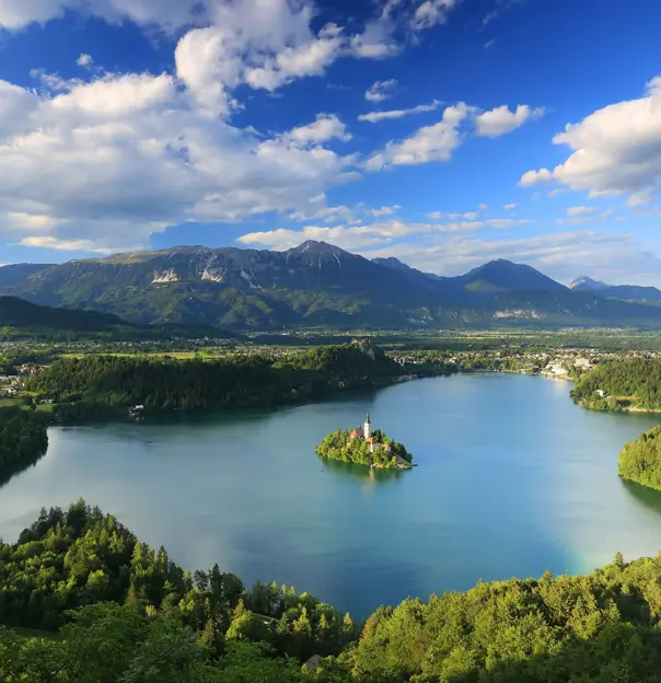  Wide angle shot of Lake Bled, showing its forested surrounding areas, and mountains in the distance. In the middle of the lake, there is a small piece of land with trees and a towered building.