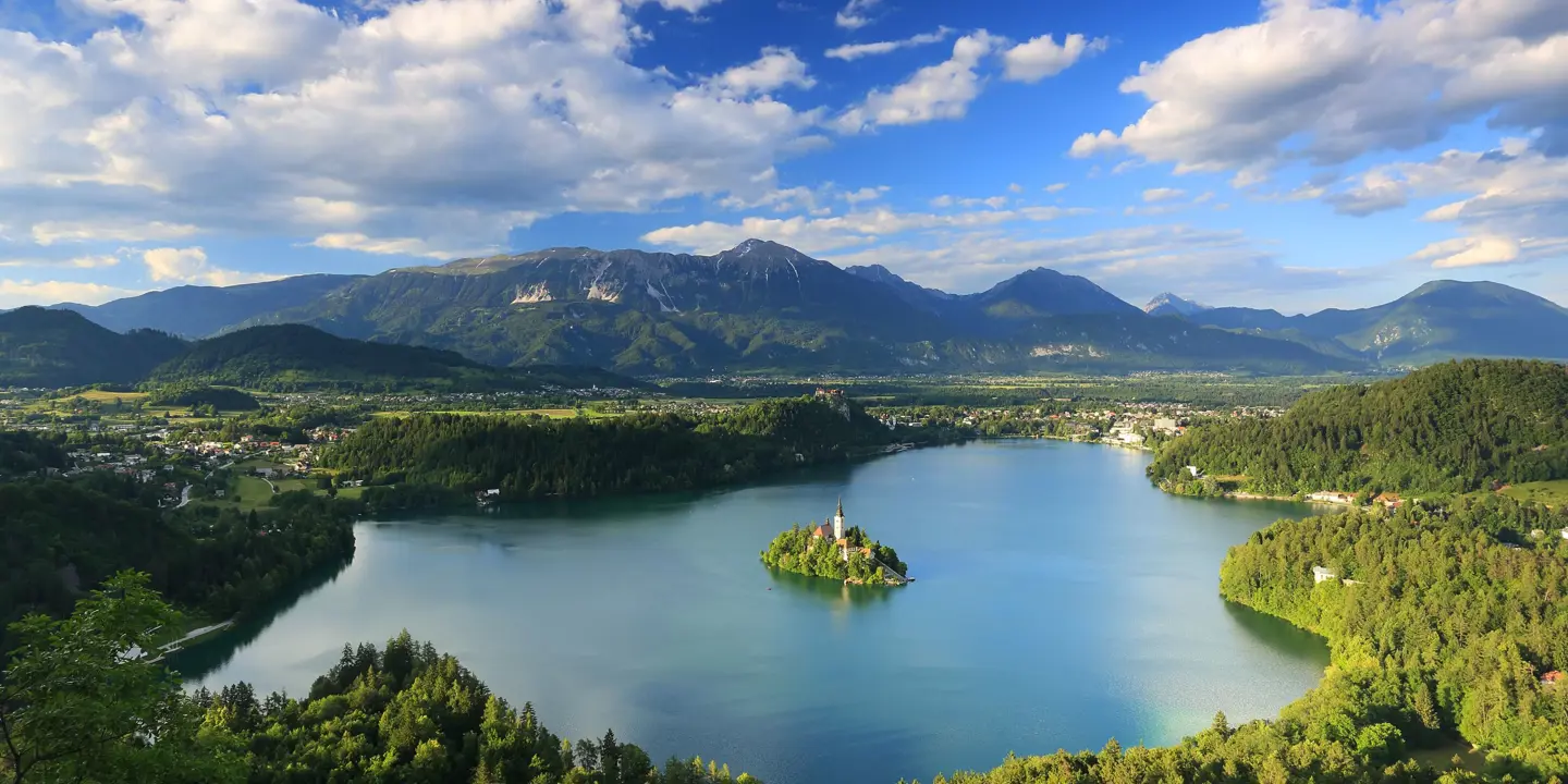  Wide angle shot of Lake Bled, showing its forested surrounding areas, and mountains in the distance. In the middle of the lake, there is a small piece of land with trees and a towered building.