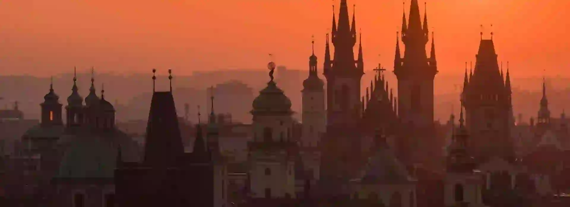 Prague cityscape against an orange sky. Silhouettes of tall buildings with spiked turrets.