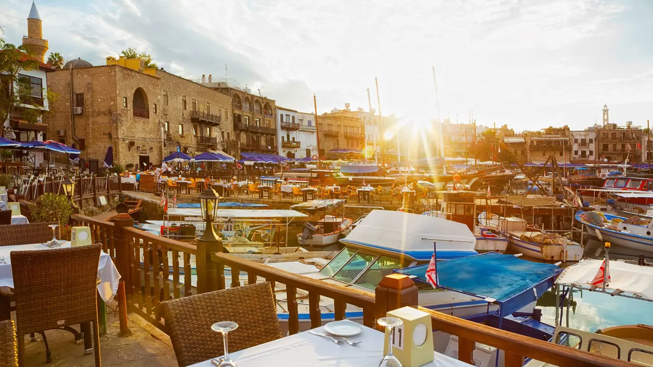 View of a marina from a restaurant, with a laid table in the forefront behind a wooden barrier dividing the boats on the water and the land. Other buildings and restaurants can be seen on the other side, with blue umbrellas and outdoor seating. The sun is poking over the buildings, with beams shining into the camera