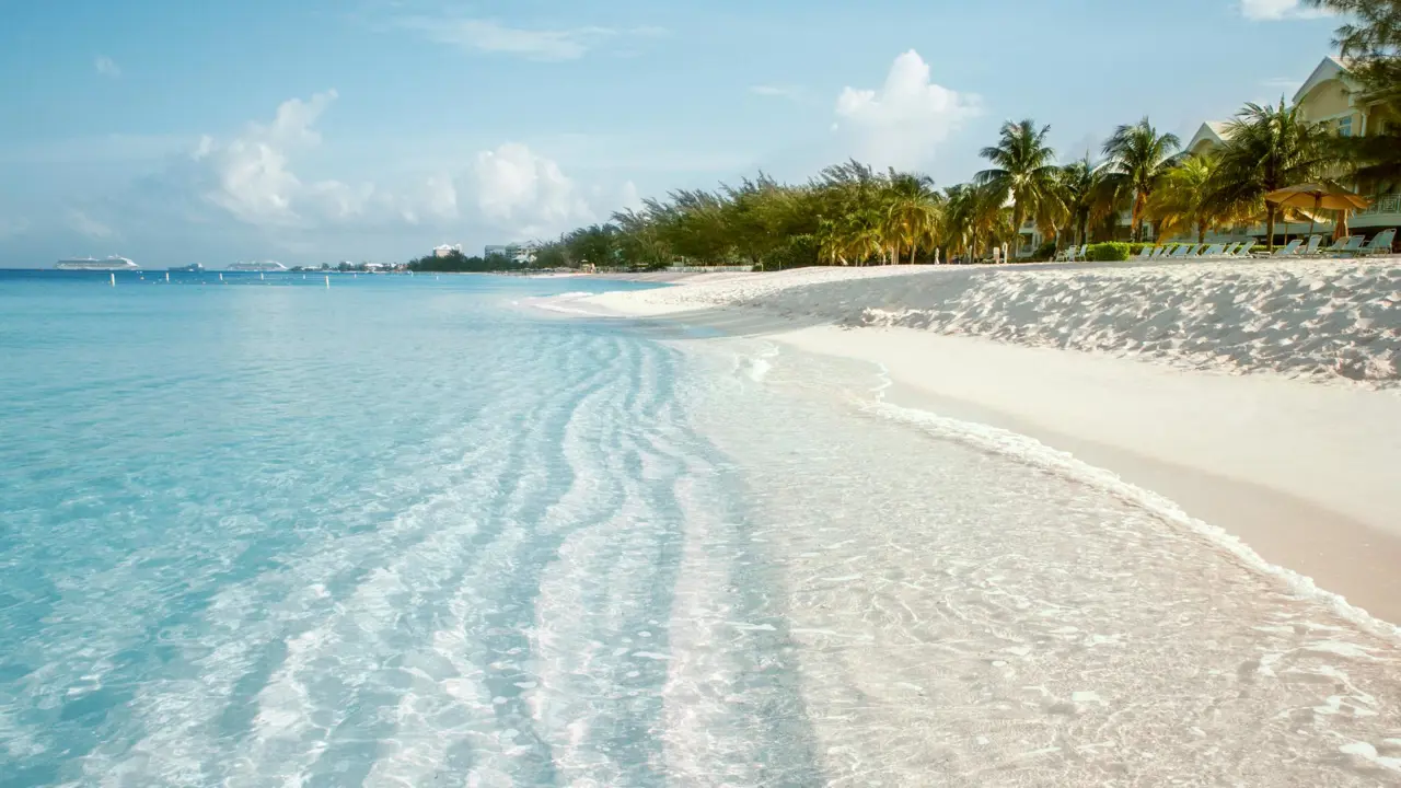 The pristine white sands and crystal clear shallow waters of Seven Mile Beach in Grand Cayman, with palm trees and resorts lining the shore under a soft, hazy blue sky