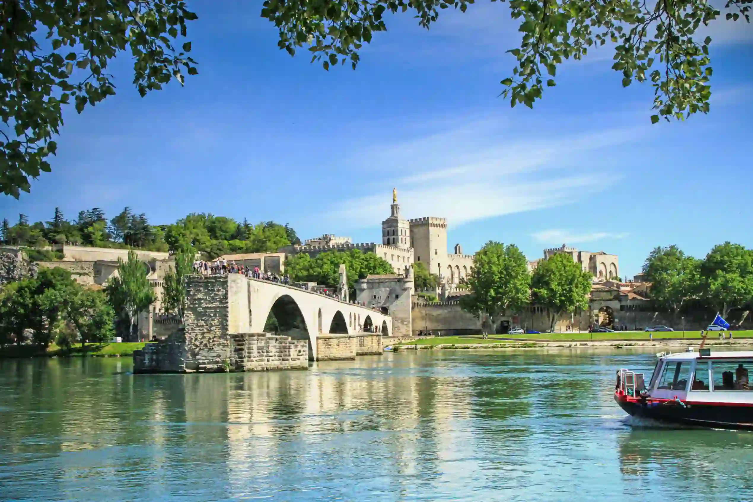 View of the Pont D’Avignon bridge, with the river water in the forefront and a boat coming from the right. In the distance, on the other side of the bridge is a grey palace. The sky is blue.