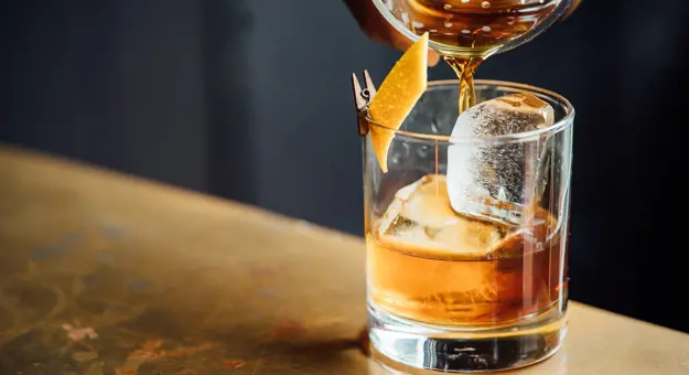 Close up on a whisky being poured, with ice in the glass and lemon peel clipped to the side of the glass. 