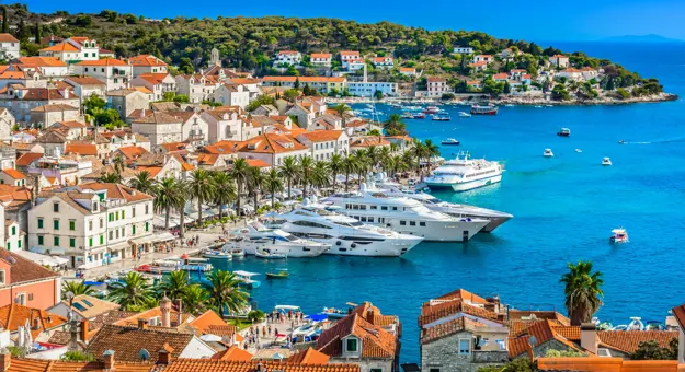 High angle shot of a town on the waterfront, with yachts docked on the water. Rows of palm trees all round the water's edge, in front of cream houses with orange roofs, and orange roofs in the forefront. Further away, land covered in trees and a blue sky.