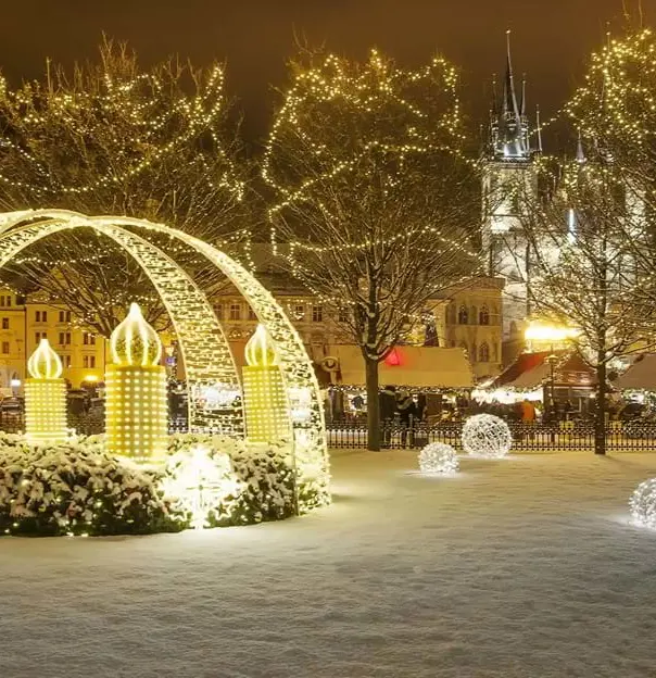 Crossing archways of fairylights with giant lit up artificial candles, fairy lit trees and balls of fairy lights on the snowy grass. A gothic church behind the trees in the distance and a lamp post in the right forefront.