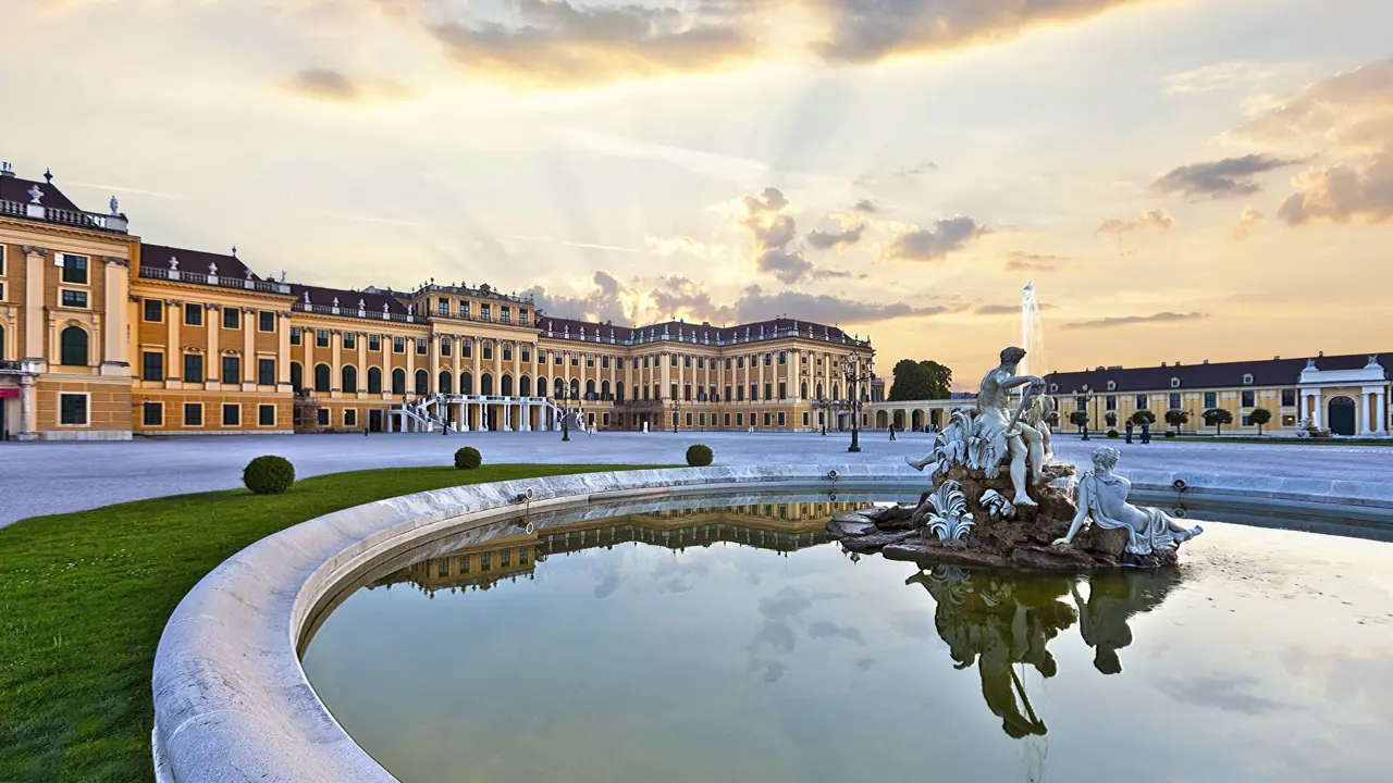 A pond with a fountain with a white statue of people sitting on a rock in the right forefront, behind is Schoenbrunn Palace, a yellow and orange, wide building in front of a bright sky with a yellow hue.