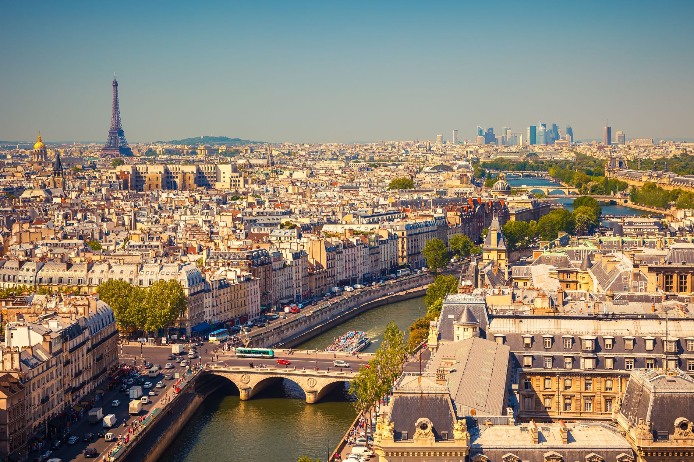 High angle view of the city of Paris, with a bridge and a main road with cars in the forefront. View of all the buildings of the city, with the Eiffel tower poking out in the distance to the left. To the right, behind the river, the faint view of skyscrapers in the distance.