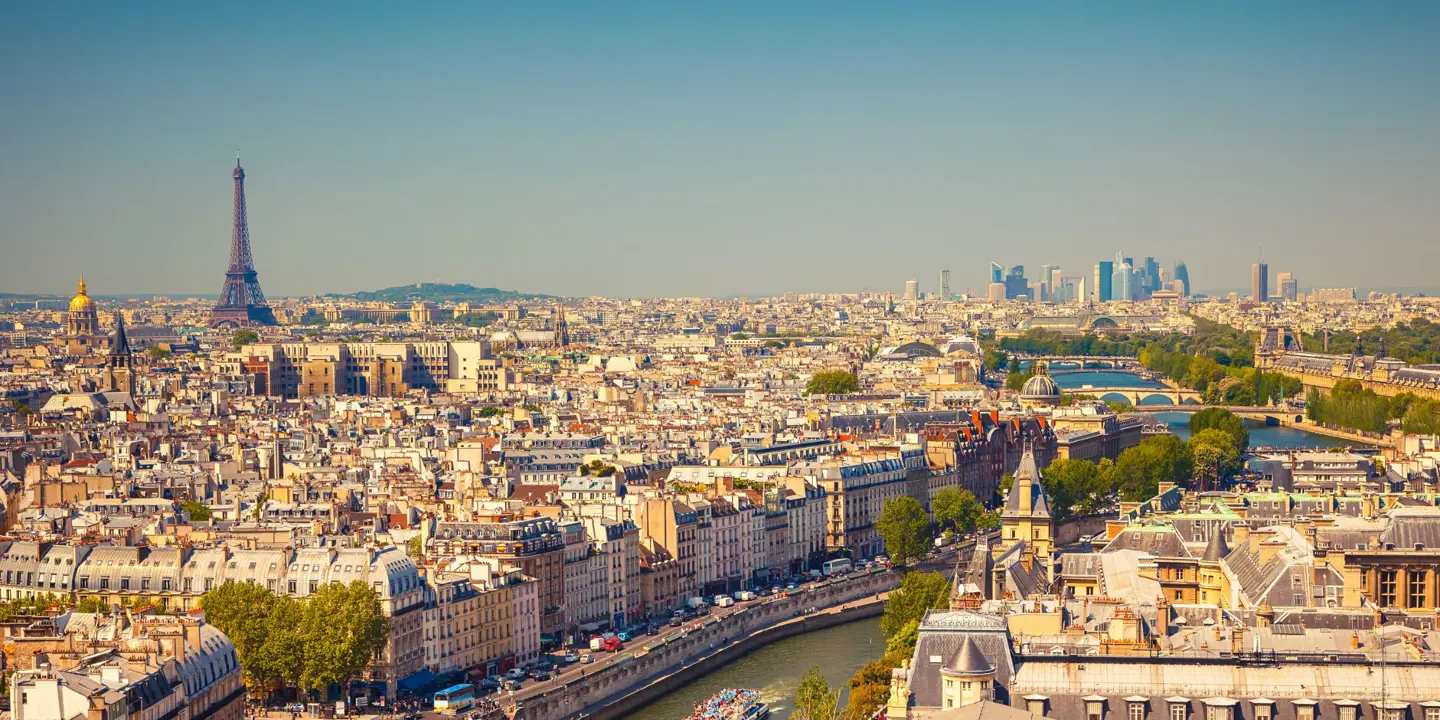 High angle view of the city of Paris, with a bridge and a main road with cars in the forefront. View of all the buildings of the city, with the Eiffel tower poking out in the distance to the left. To the right, behind the river, the faint view of skyscrapers in the distance.