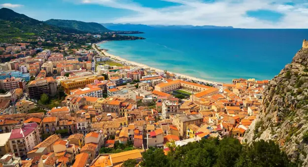 A panoramic view of Cefalù, Sicily, featuring its historic seaside town with terracotta rooftops, a sandy beach, and the rugged coastline beneath a lightly cloudy sky