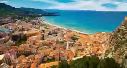 A panoramic view of Cefalù, Sicily, featuring its historic seaside town with terracotta rooftops, a sandy beach, and the rugged coastline beneath a lightly cloudy sky