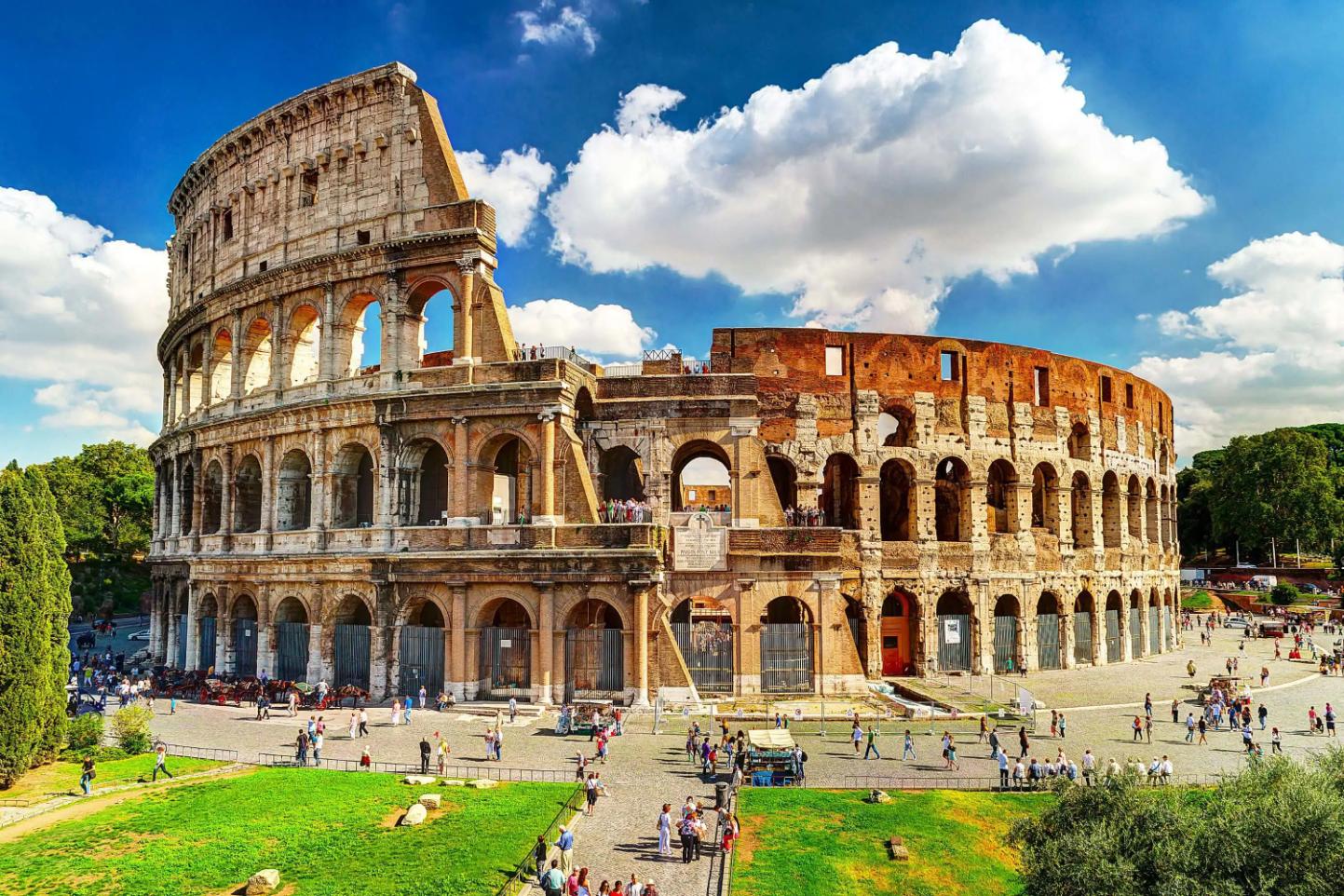 The Colosseum in Rome, a vast ancient Roman amphitheatre with partially preserved stone arches and tiered seating, set against a blue sky with scattered clouds