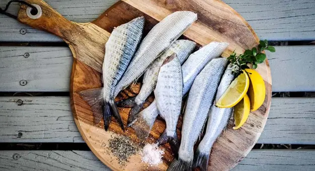 Whole silver fish on a wooden platter, with lemon slices, herbs and piles of salt & pepper next to them