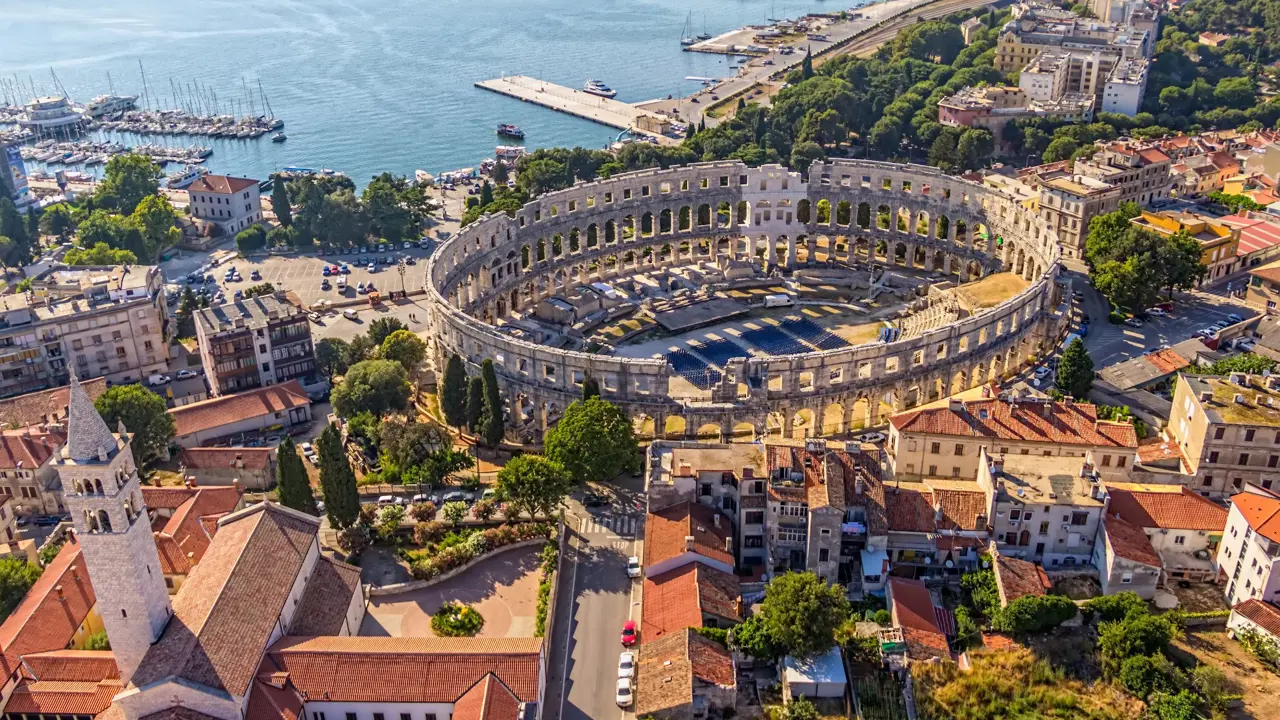 Bird's eye view of the ruins of an Amphitheatre and its surrounding area. A church with a tower in the left forefront, and regular buildings to the right. A boat harbour to the top right of the image, next to the rest of the sea to the right. 