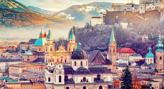 Shot of Salzburg, showing the top half of the city's buildings, with turquoise turrets dotted around the image and a wide church in the forefront and a cathedral behind it. Mountainous landscape behind and the fortress on the hill to the right.