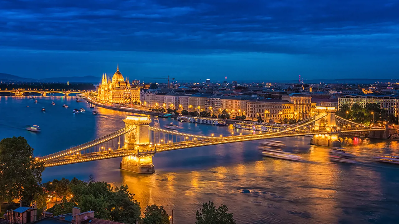 High angle shot of Széchenyi Chain Bridge, going over the Danube River at night time. With a view of the Pest side, the Parliament building in the distance