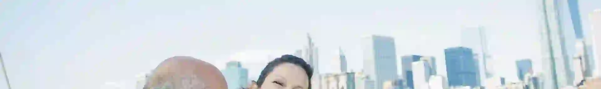 Two people smiling happily on a boat in New York City, with the city skyline visible in the background