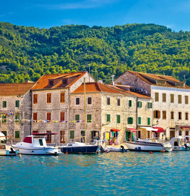 View of bricked buildings on the waterfront from the sea, with boats docked in front of them. Along the street, on the left, multi-coloured bunting, and to the right, sun roofs pulled out from the buildings. Above, mountains covered in trees and above that, a blue sky.