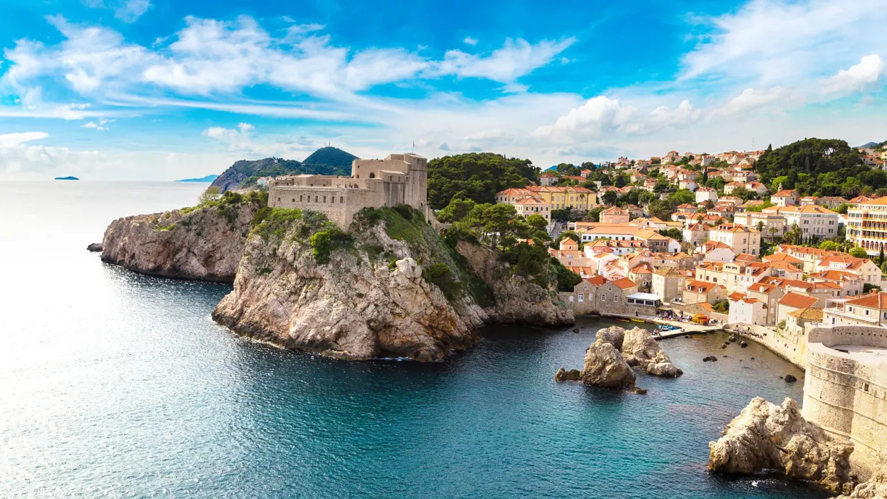 View of a coastal town with a large stone building on top of a rocky hill, above the sea. The buildings in the town on the right are mainly white and cream coloured and have orange roofs. The sky is blue but slightly cloudy.