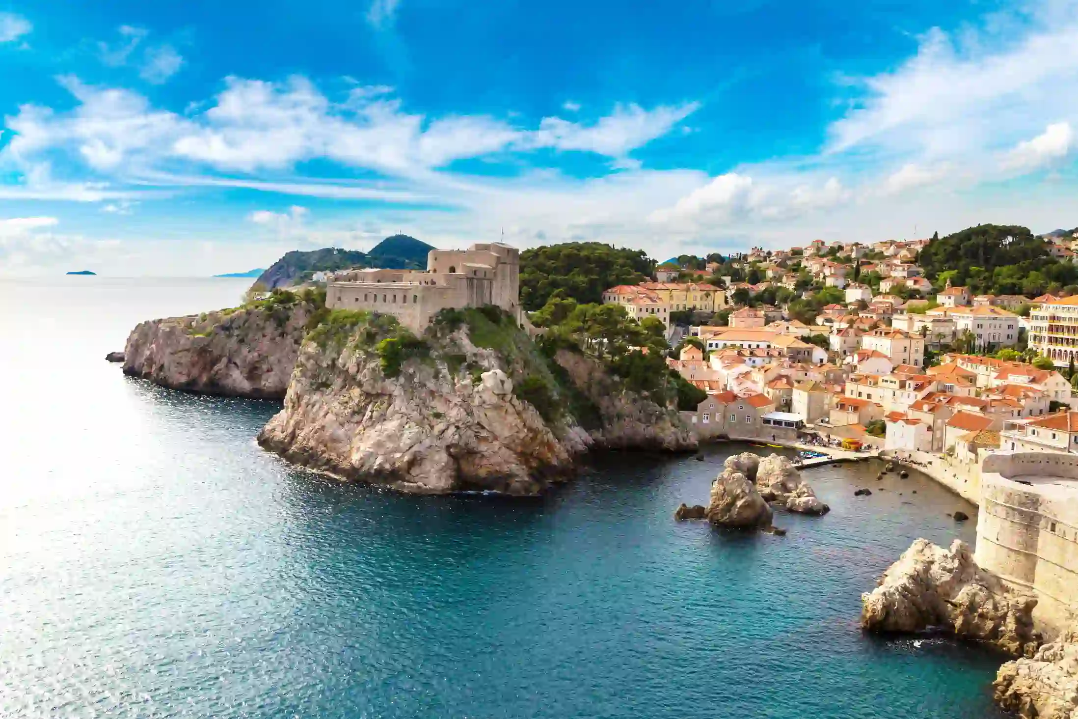 View of a coastal town with a large stone building on top of a rocky hill, above the sea. The buildings in the town on the right are mainly white and cream coloured and have orange roofs. The sky is blue but slightly cloudy.