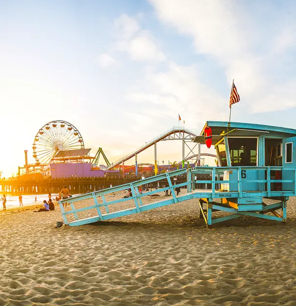 A teal blue lifeguard hut on Santa Monica Beach, with the iconic pier and Ferris wheel in the background as the sun goes down