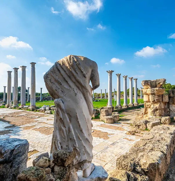 View of Roman ruins, with the back of a headless marble statue of a person. Further away, there is a green lawn surrounded by marble pillars. 