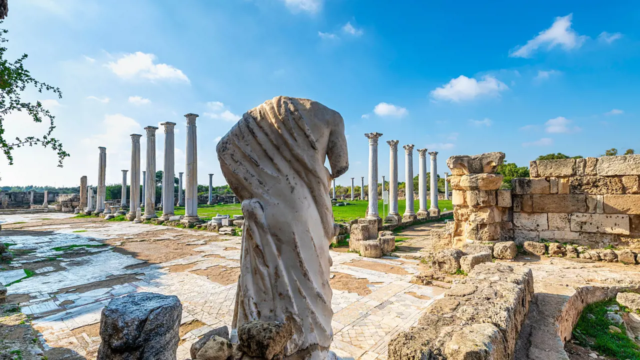 View of Roman ruins, with the back of a headless marble statue of a person. Further away, there is a green lawn surrounded by marble pillars. 