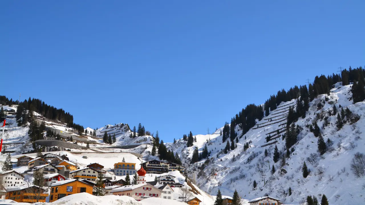 Low-angle view of St Anton village covered in thick snow, with a few houses and hotels going up the mountain and fir trees dotted all over. In front of a blue sky.