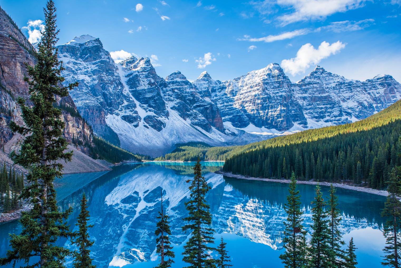 Snow-capped Rocky Mountains towering over the serene, turquoise Moraine Lake in Canada, with dense pine forests lining the shores and a clear blue sky above