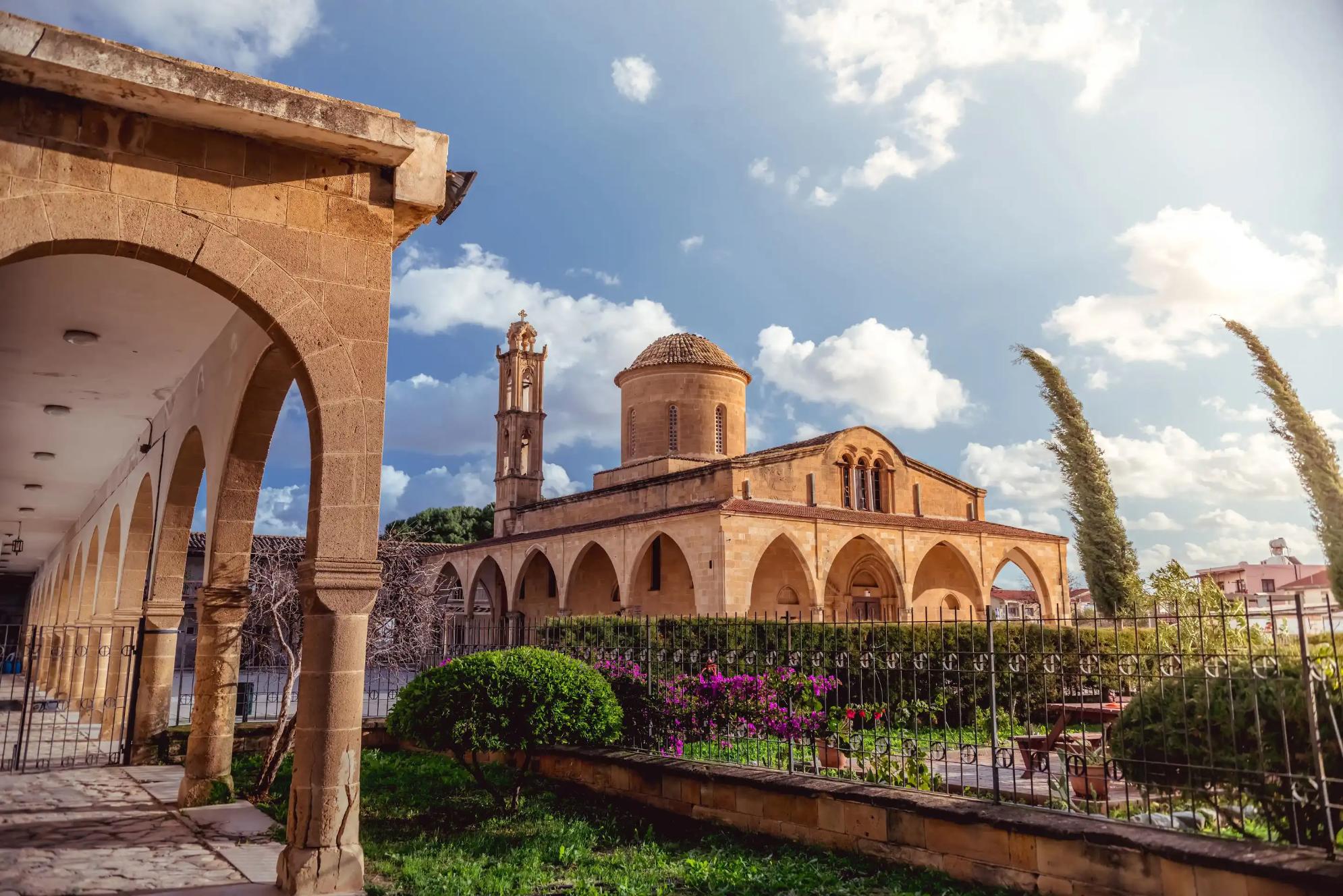A stone Greek Orthodox monastery with arched walkways, a domed roof, and a bell tower. A well-kept garden with flowers and shrubs lines the foreground under a sunny sky