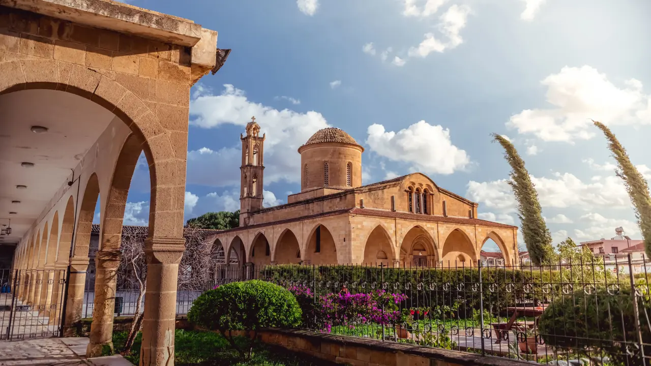 View of a beige stone building, archways round the bottom, a triangular roof, dome shaped tower, and a thin tower. In front is a gated garden with bushes, trees and flowers. To the left is a beige stone archway