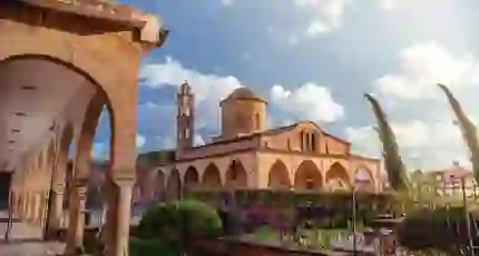 A stone Greek Orthodox monastery with arched walkways, a domed roof, and a bell tower. A well-kept garden with flowers and shrubs lines the foreground under a sunny sky