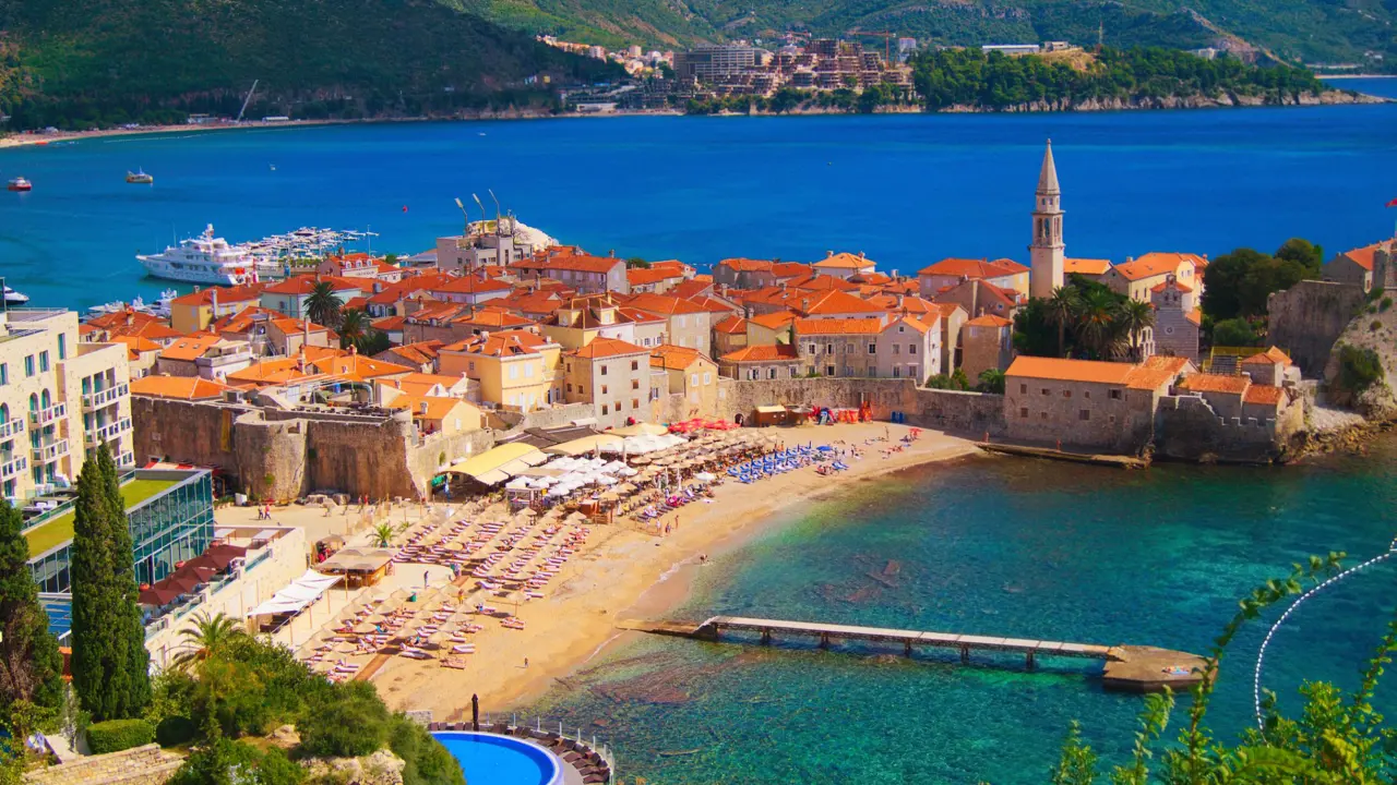  High angle view of a small beach, with sunloungers laid out and a turquoise sea. A group of buildings with orange roofs behind the beach and a bright blue sea behind there, with the start of a mountain behind it.