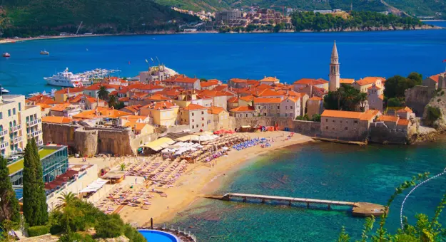 High angle view of a small beach, with sunloungers laid out and a turquoise sea. A group of buildings with orange roofs behind the beach and a bright blue sea behind there, with the start of a mountain behind it.