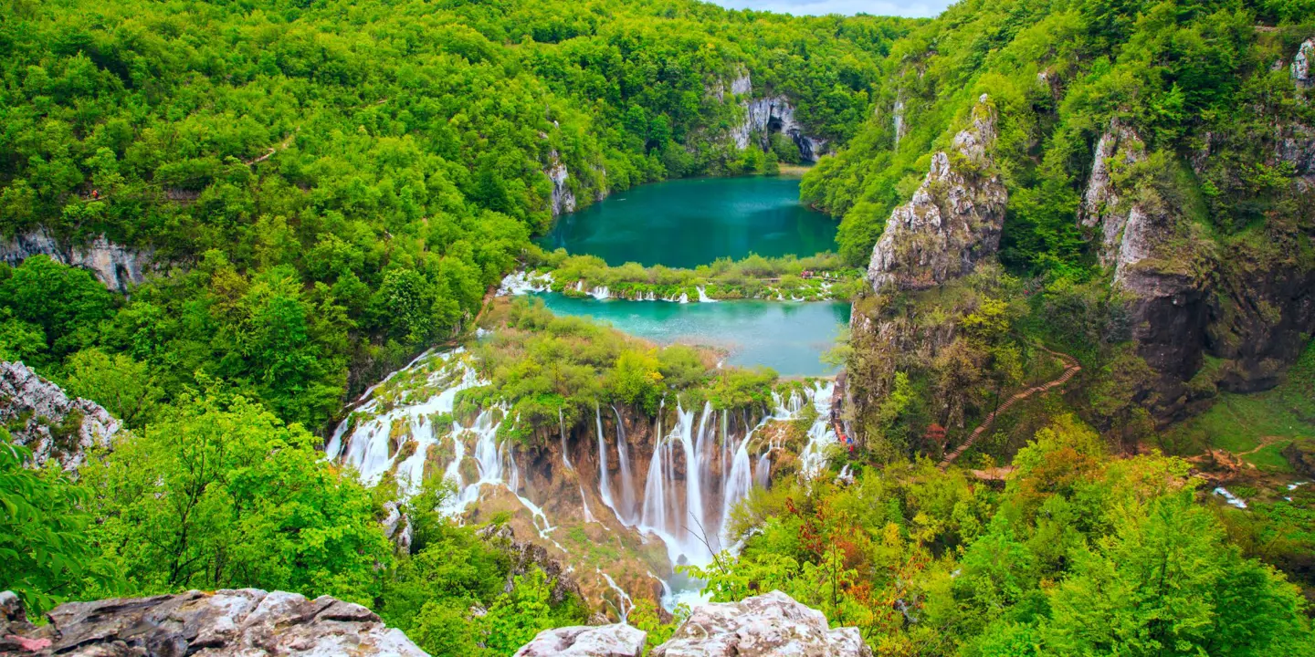 High shot of waterfalls in front of a dark turquoise body of water, surrounded by forested hills. In the forefront is a piece of rock and trees.