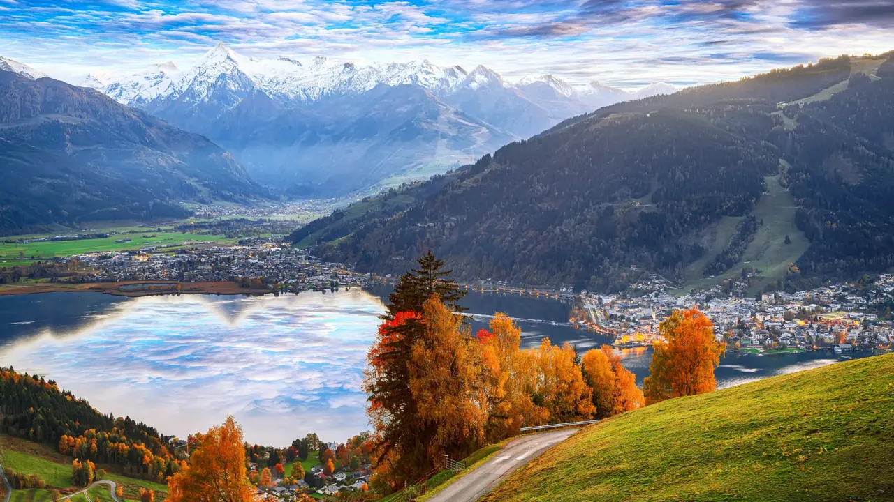 View of Austrian mountains, with some in the distance with snow on the top. Below is a town where small buildings can be seen, behind a lake. To the right another town, and in the forefront, orange autumnal trees and a grassy hill and a pathway