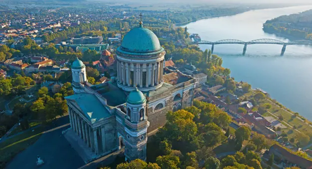 Bird's eye view of the Basilica of Esztergom, with it's turquoise domed roof with a cross on in the centre, and two smaller towers at the front with domed turquoise tops. Danube river in the background, with a bridge going across.