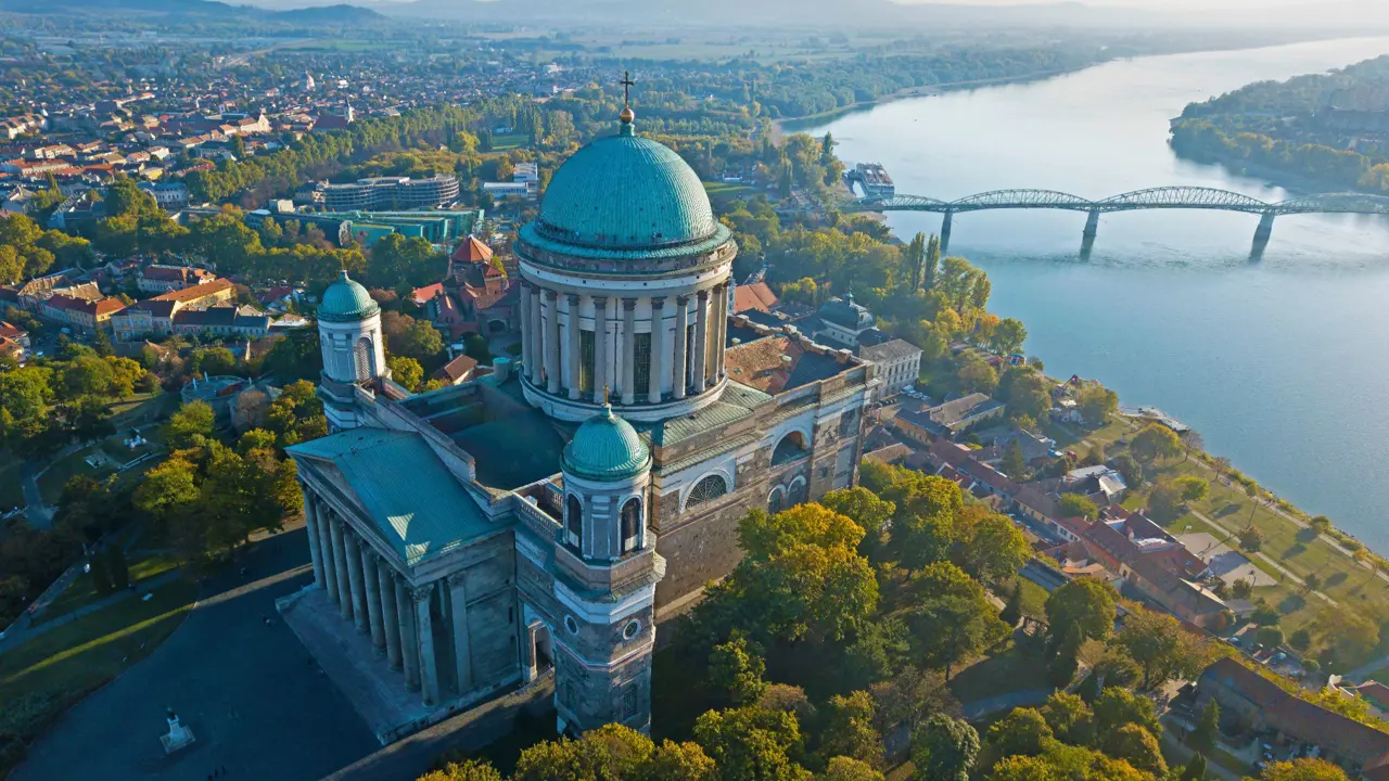 Bird's eye view of the Basilica of Esztergom, with it's turquoise domed roof with a cross on in the centre, and two smaller towers at the front with domed turquoise tops. Danube river in the background, with a bridge going across.