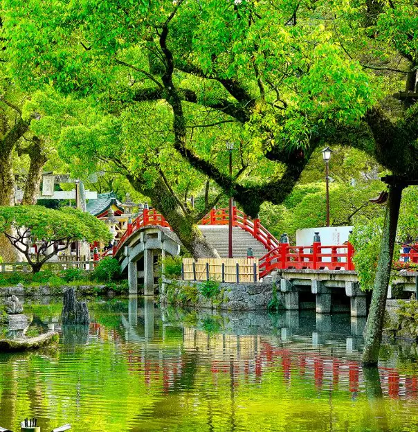 A serene Japanese garden with a red arched bridge over a reflective pond, surrounded by lush green trees at Dazaifu Tenmangu Shrine in Fukuoka