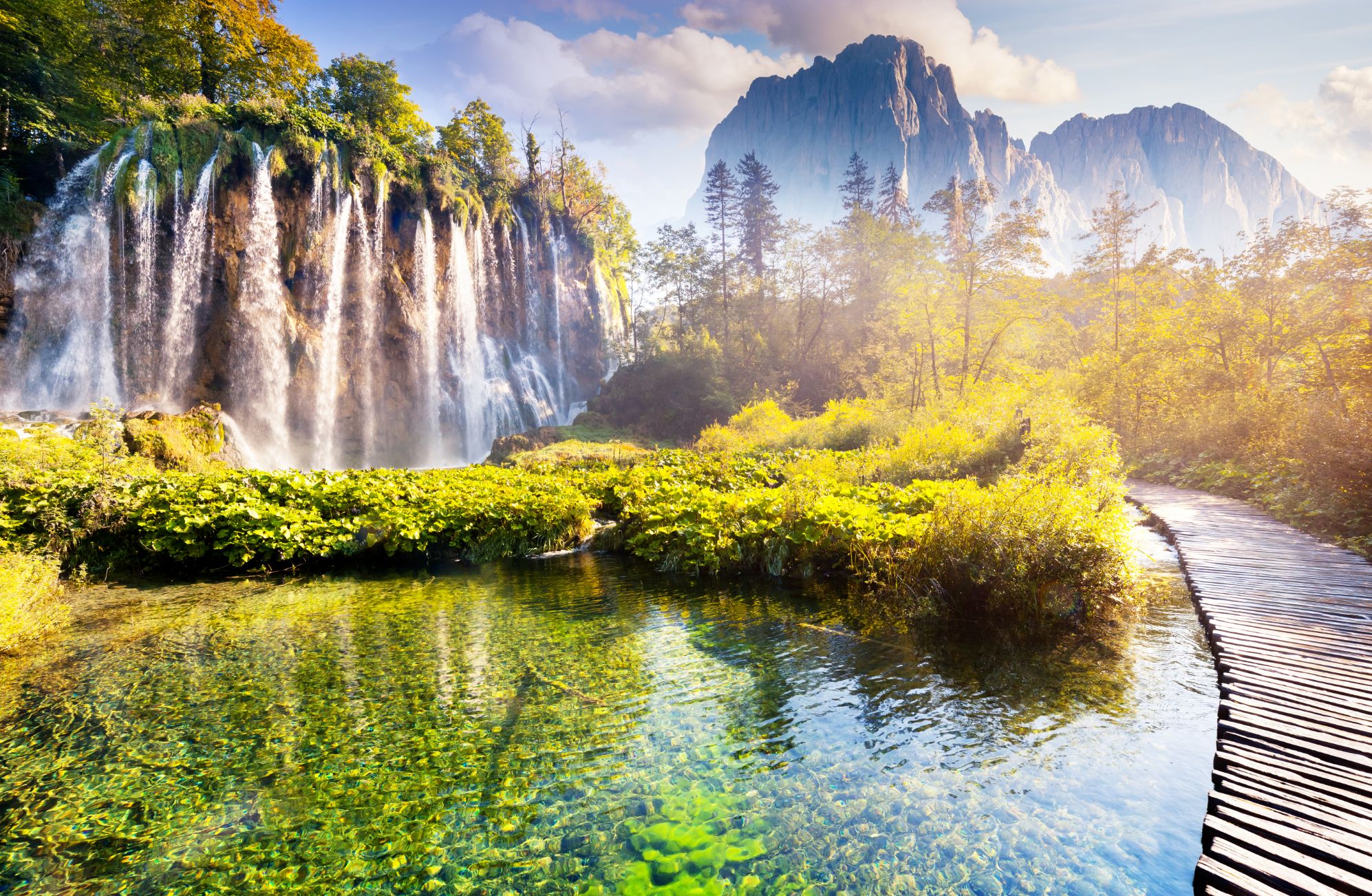 Shot of the Plitvice Lakes, showing a waterfall with bushes at the bottom, next to a small body of water. On the right, there is a path curving to the left and mountains in the distance.