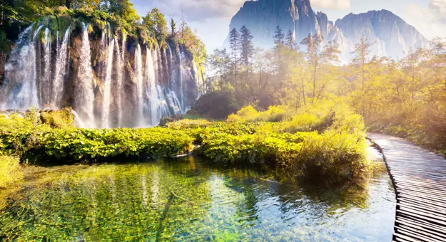 Shot of the Plitvice Lakes, showing a waterfall with bushes at the bottom, next to a small body of water. On the right, there is a path curving to the left and mountains in the distance.