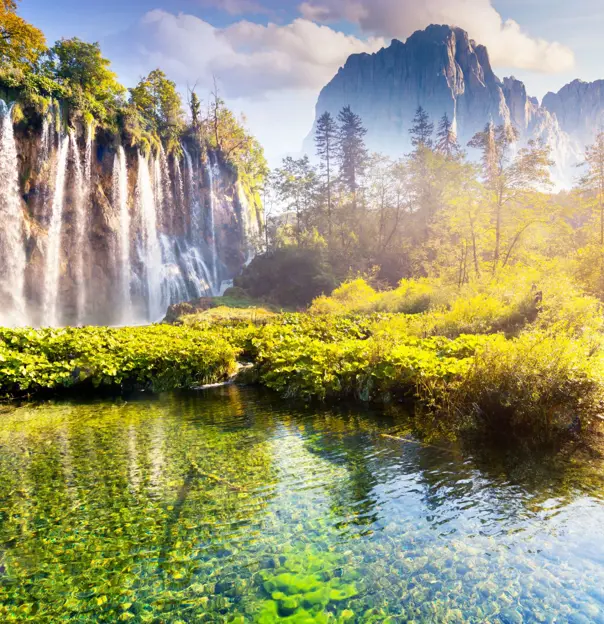 Shot of the Plitvice Lakes, showing a waterfall with bushes at the bottom, next to a small body of water. On the right, there is a path curving to the left and mountains in the distance.