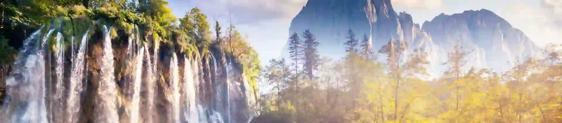 Shot of the Plitvice Lakes, showing a waterfall with bushes at the bottom, next to a small body of water. On the right, there is a path curving to the left and mountains in the distance.