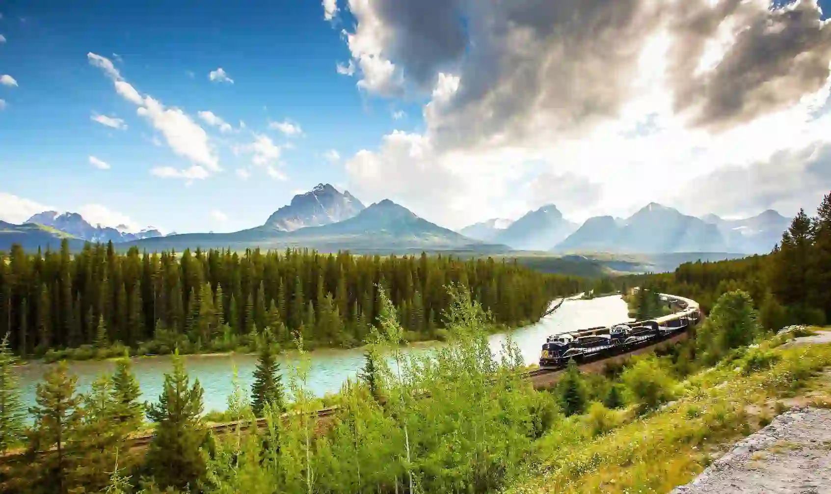 A train travelling through the Canadian Rockies, with rugged mountains in the background and dense evergreen forests lining the tracks