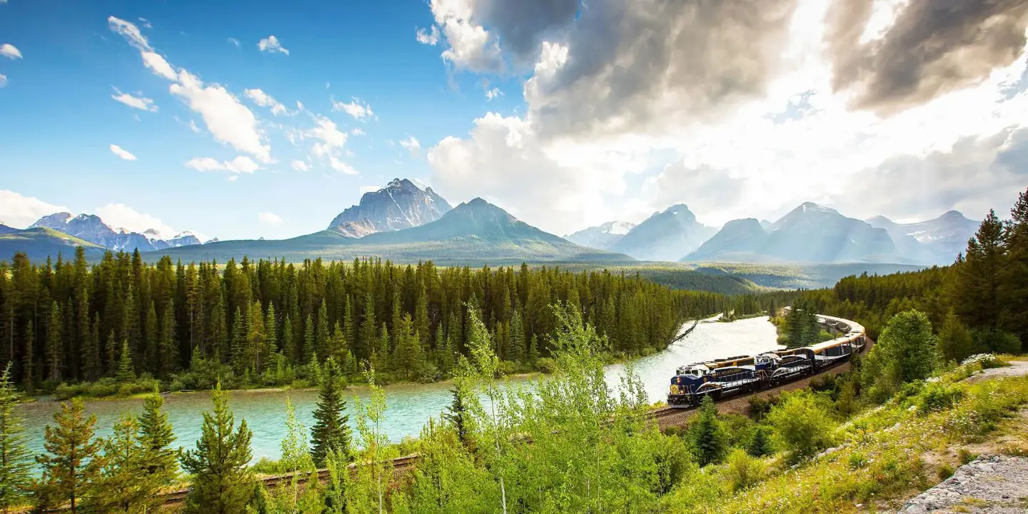 A train travelling through the Canadian Rockies, with rugged mountains in the background and dense evergreen forests lining the tracks