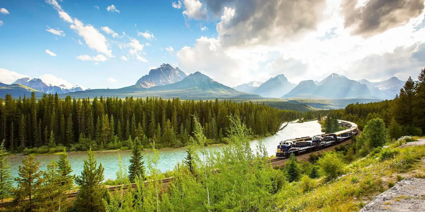 The Rocky Mountaineer train travelling through the Canadian Rockies, with rugged mountains in the background and dense evergreen forests lining the tracks