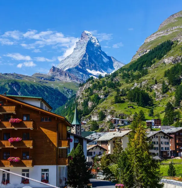 Daytime view of the towering Matterhorn in Zermatt, Switzerland, with traditional Swiss buildings in the foreground and green hills framing the scene