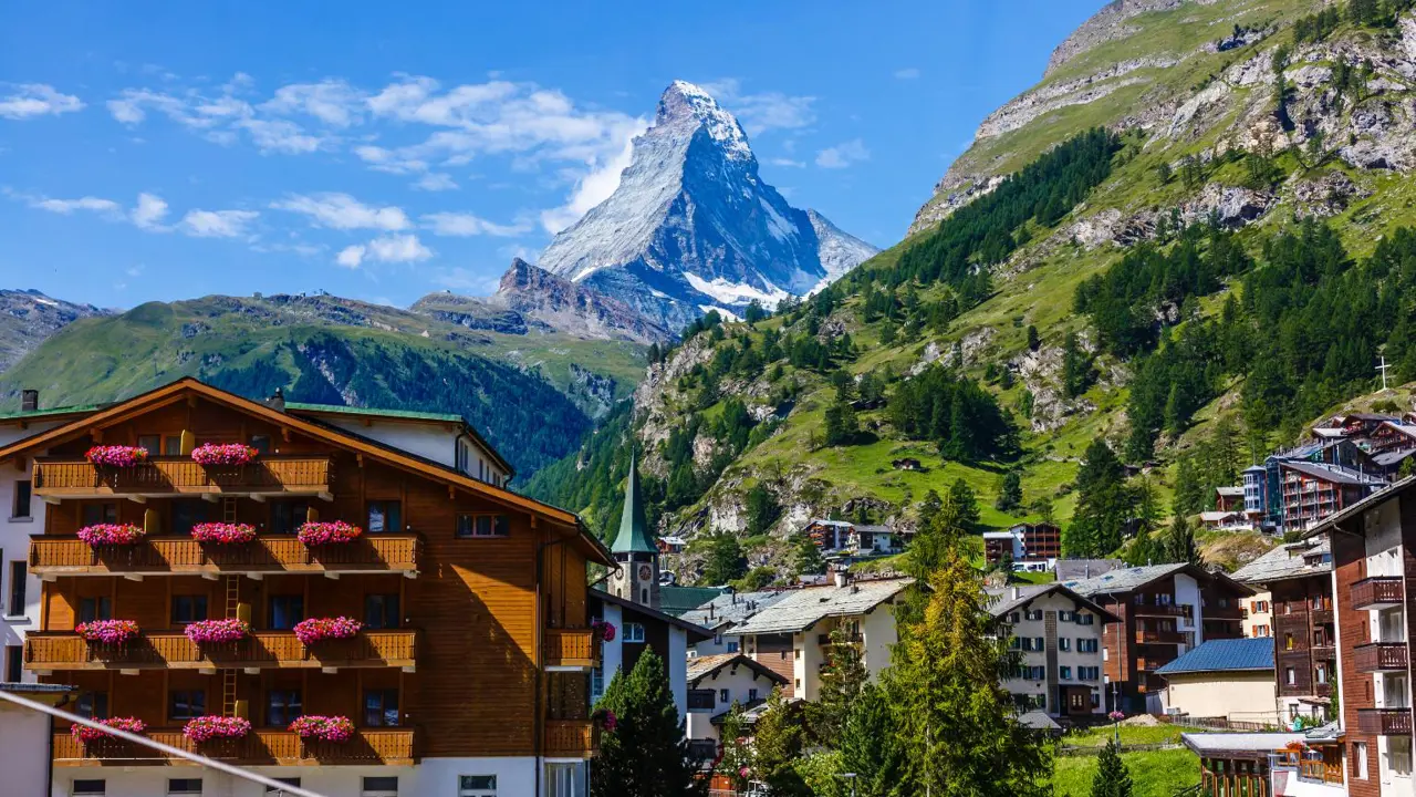 Daytime view of the towering Matterhorn in Zermatt, Switzerland, with traditional Swiss buildings in the foreground and green hills framing the scene
