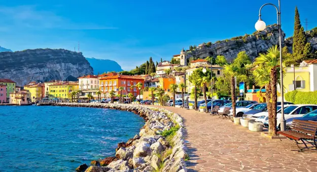 The waterfront in Torbole, Lago di Garda, featuring a paved promenade lined with colourful buildings and distant mountains rising beyond the lake